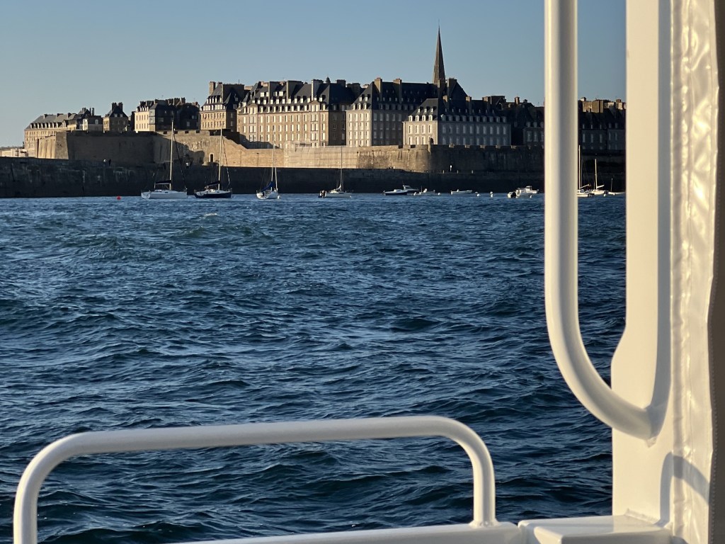 View of a coastal town with historic buildings and a distant spire from a boat on the water.