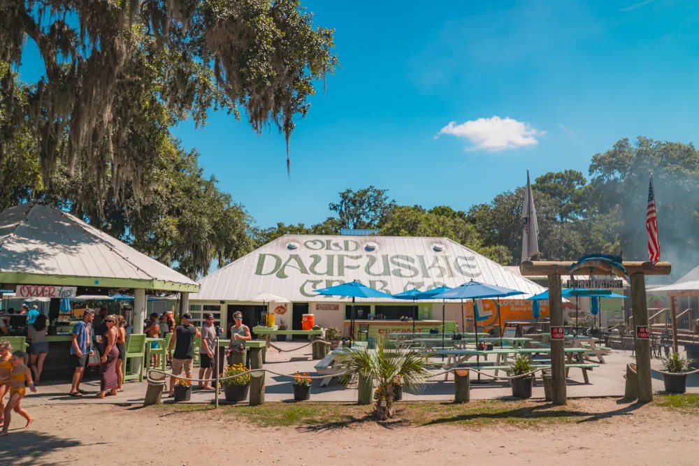 Outdoor market with tents and tables under trees, sign reads 'Old Daufuskie'.