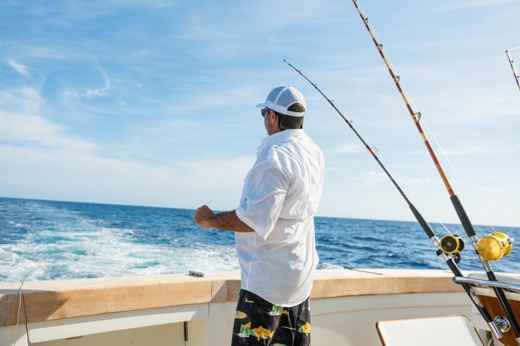 Man in shorts and cap fishing on a boat, looking at the ocean with fishing rods.