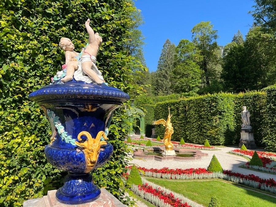 Garden with a blue urn, statues, hedges, and red flowers under a clear blue sky.