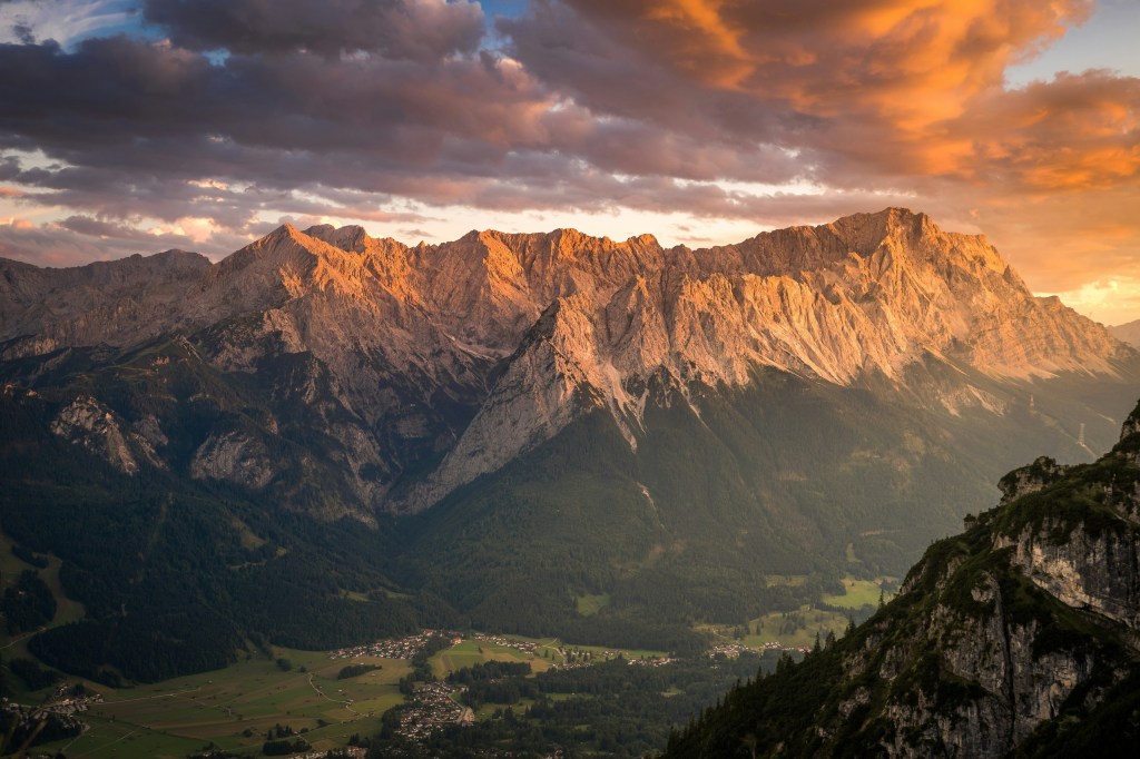 Sunlit mountain range at sunset with dramatic clouds and valley below.