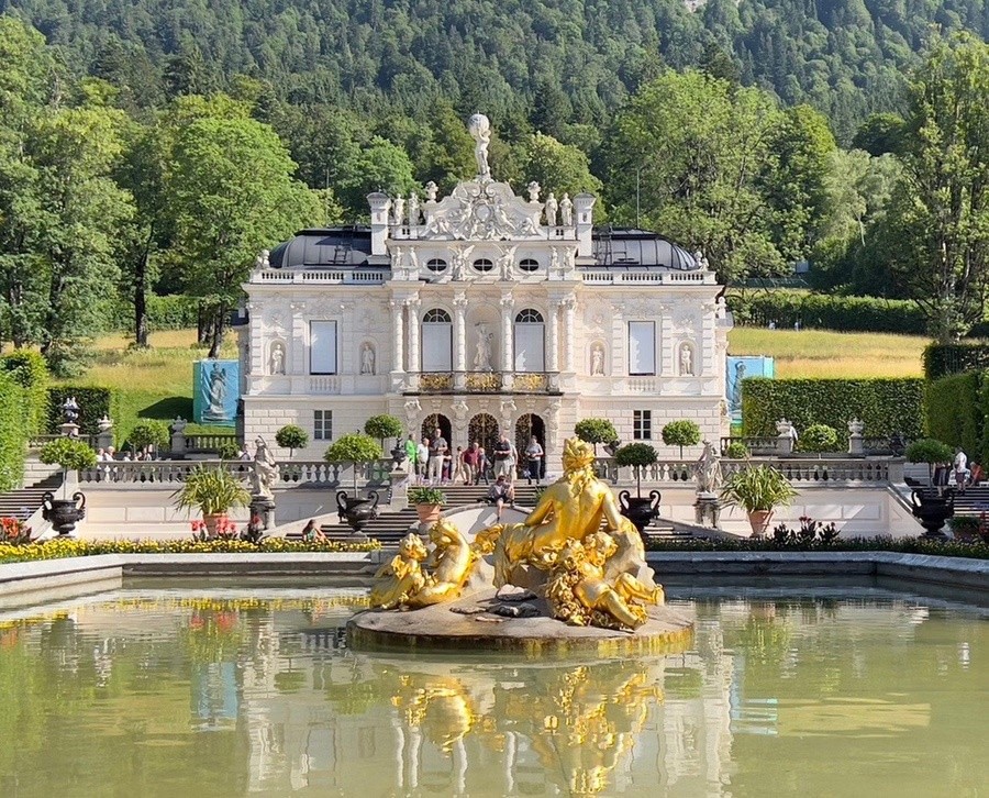 Ornate palace with white facade, golden fountain statue, and lush gardens, set against wooded hills.