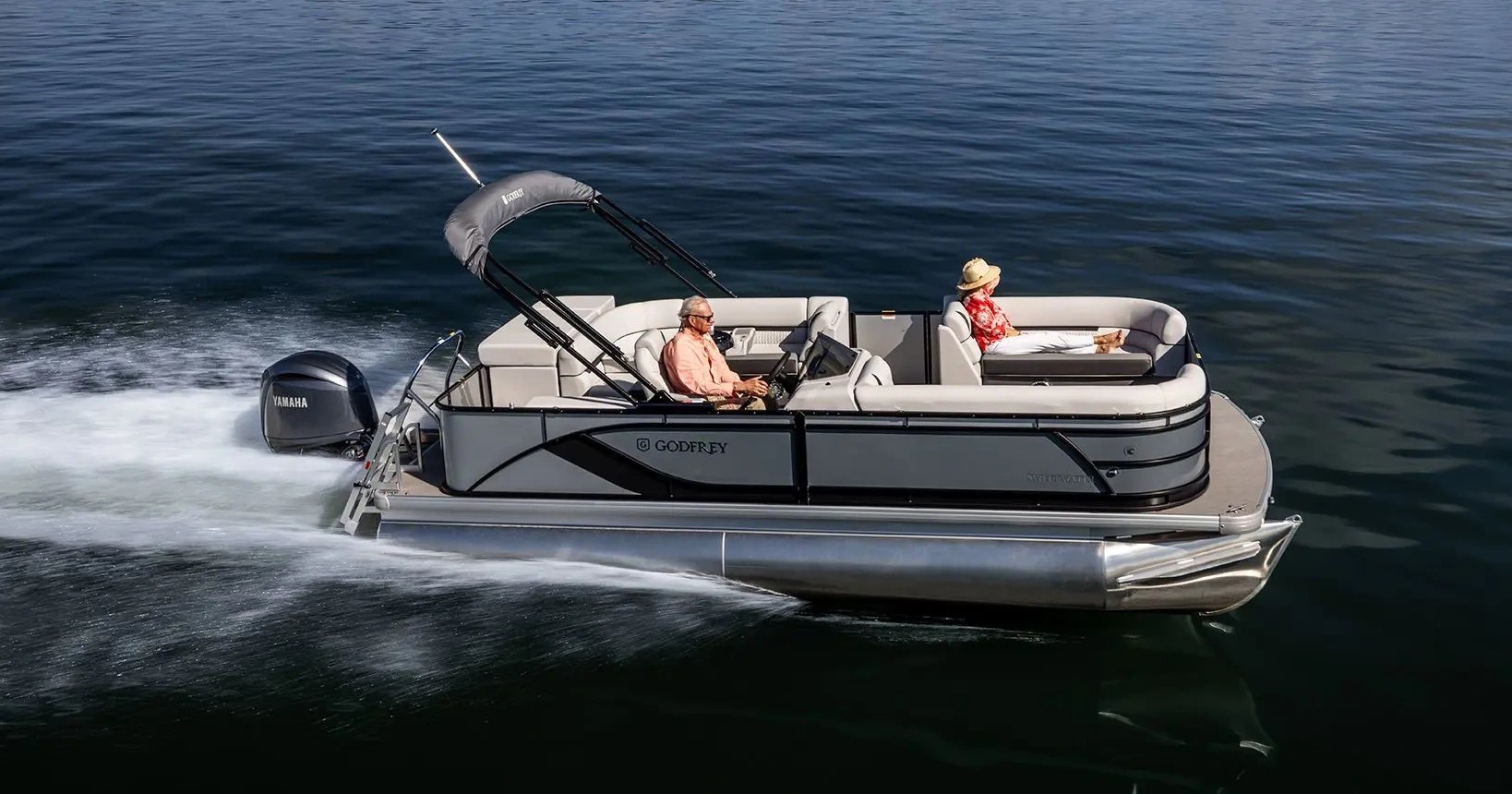 Pontoon boat with two people cruising on calm water.