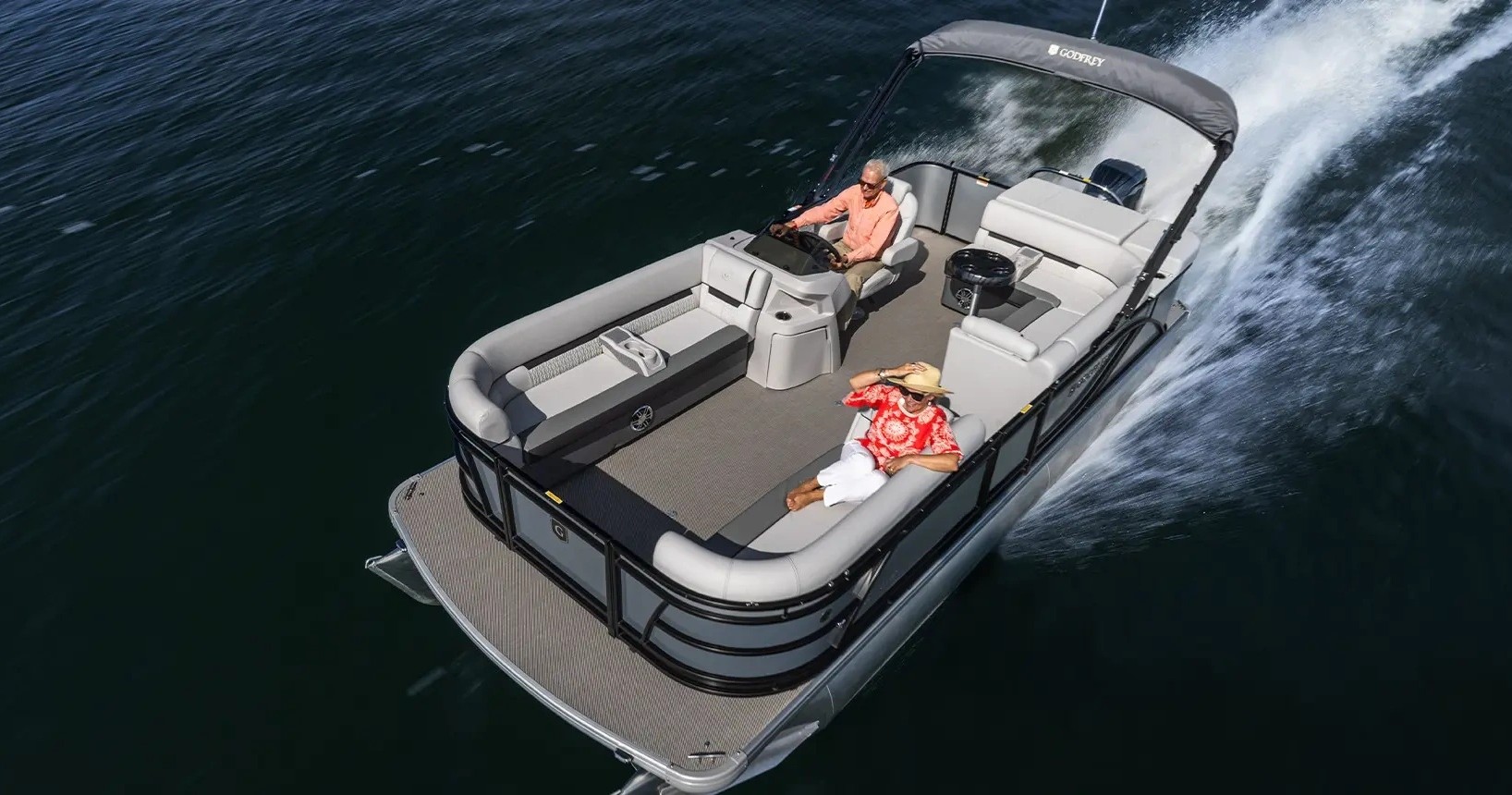 Man driving a pontoon boat with a passenger on a lake.