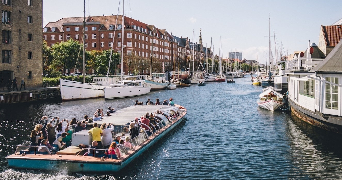 Tour boat on canal with buildings and moored boats on a sunny day.