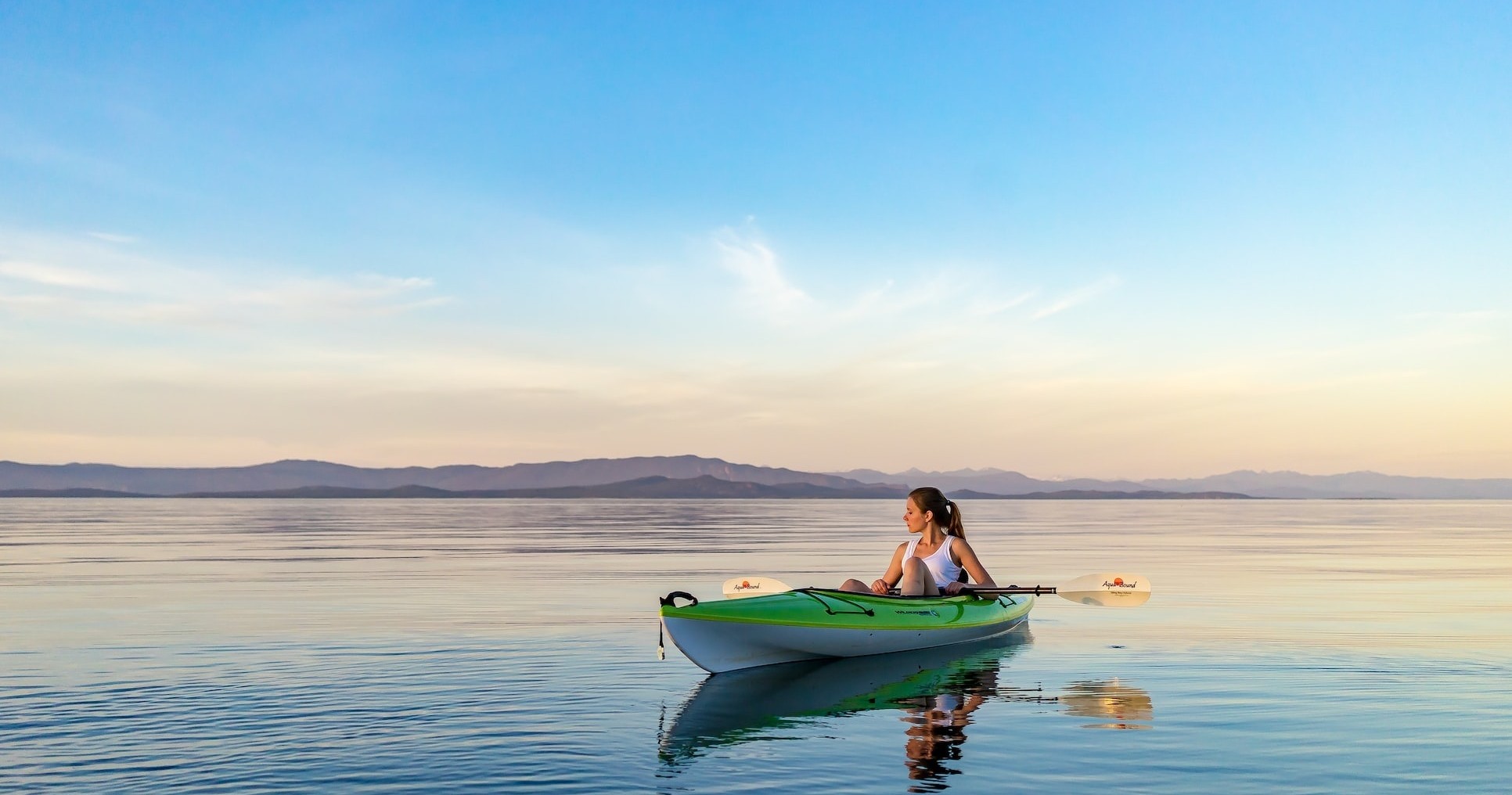 Person kayaking on calm lake at sunset with distant hills.