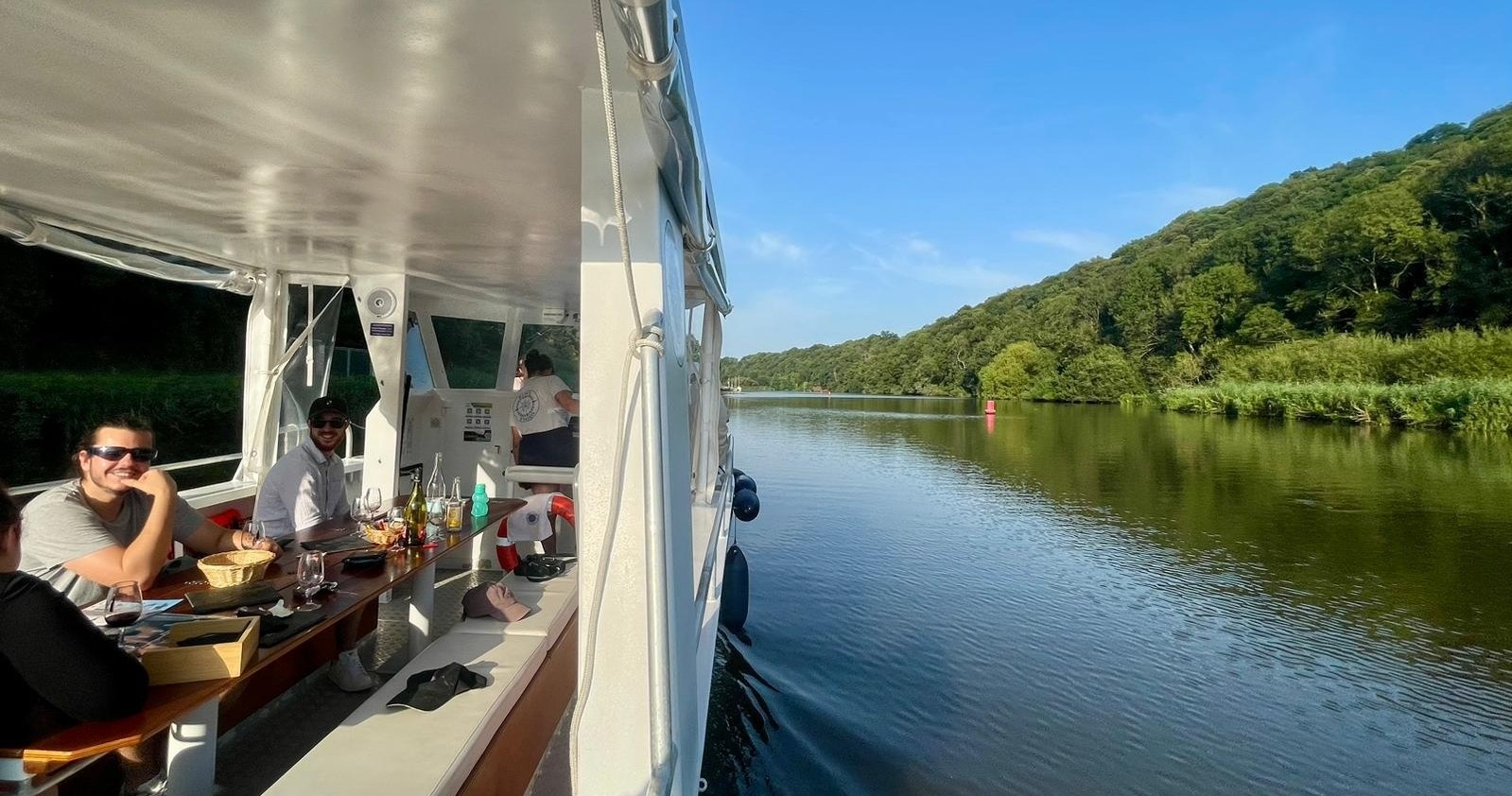 People on a boat enjoying a river cruise with greenery and blue sky.