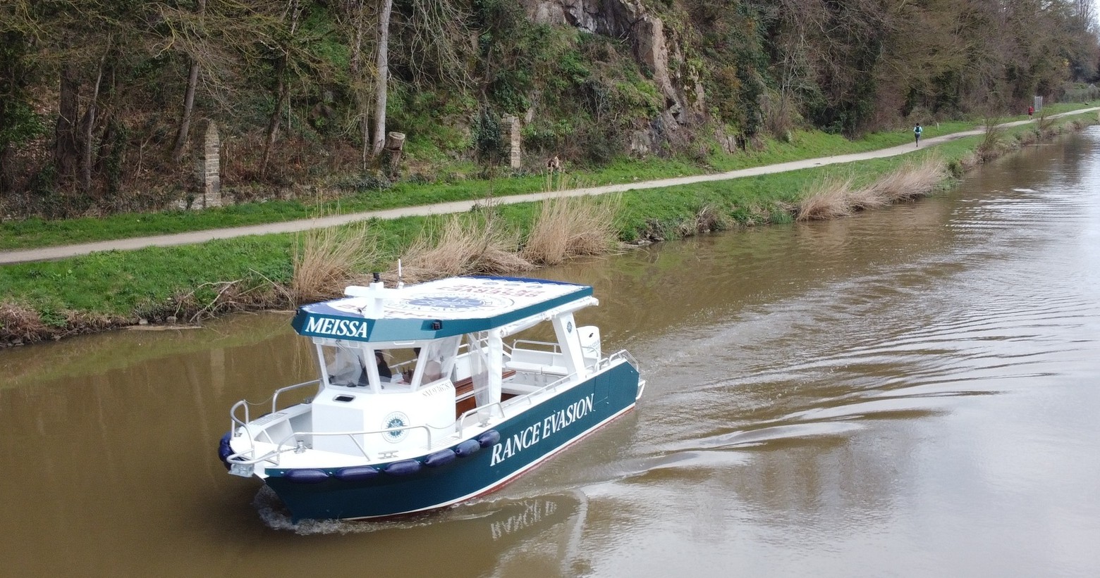 Small boat named Meissa on a river with a tree-lined path in the background.