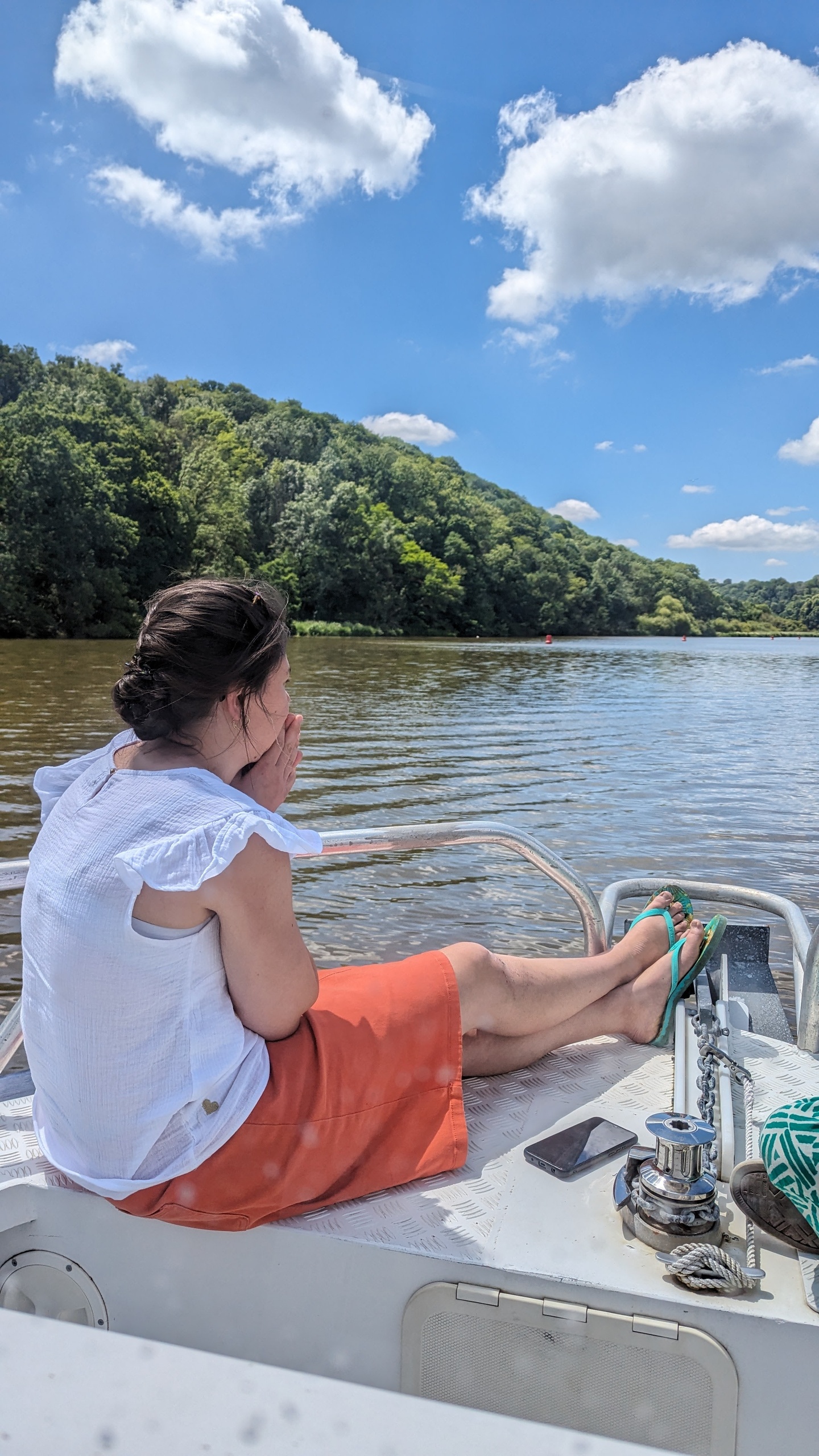 Person sitting on a boat, looking at a river and forested hills under a blue sky with clouds.