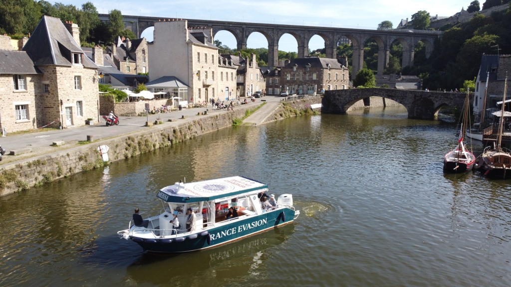 Boat on river with stone bridge and viaduct in the background, flanked by historic buildings.