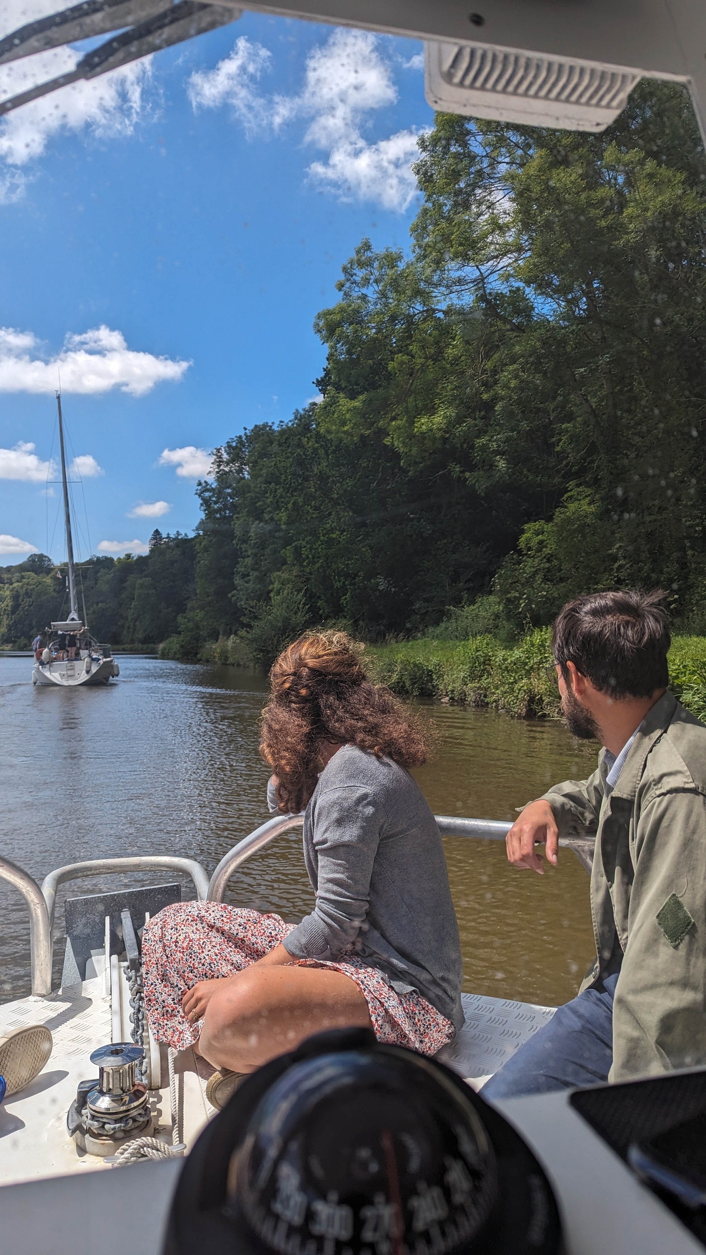 Two people on a boat watching a sailboat on a river surrounded by trees.
