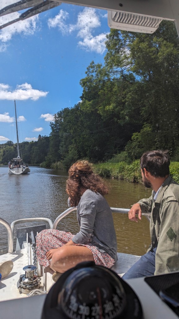 Two people on a boat watching a sailboat on a river surrounded by trees.