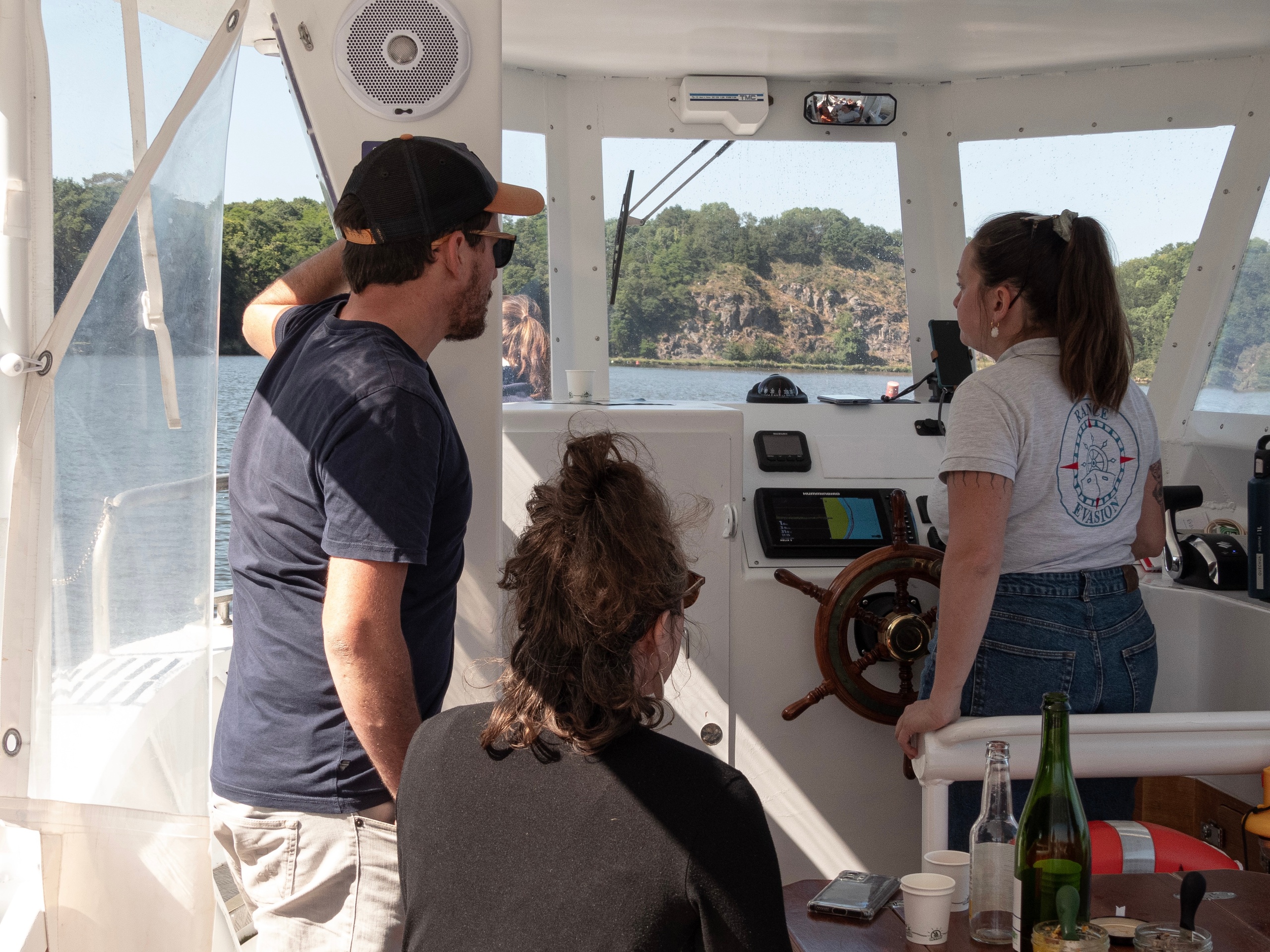 People inside a boat cockpit with water and rocky shore in view.