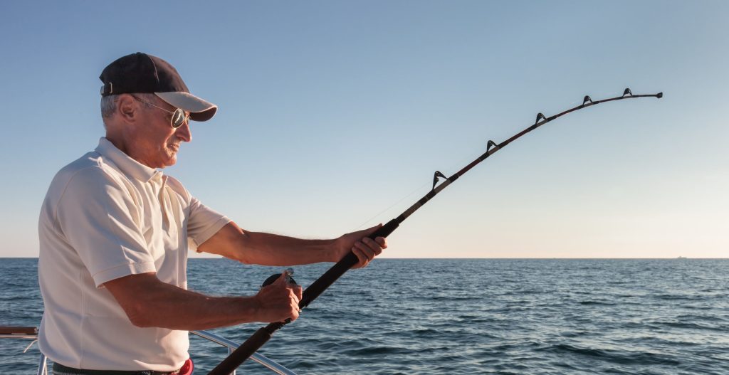 Man wearing cap fishing on a boat in open water under a clear sky.
