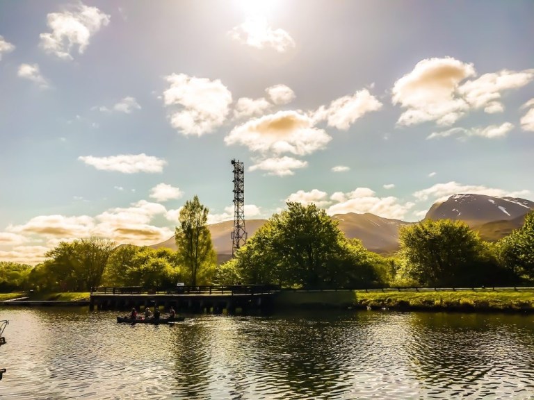 Scenic lakeside with mountains, trees, and a metal tower under a sunny sky.