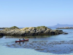 Two people in a red kayak on calm water near rocky islands, with clear blue sky.