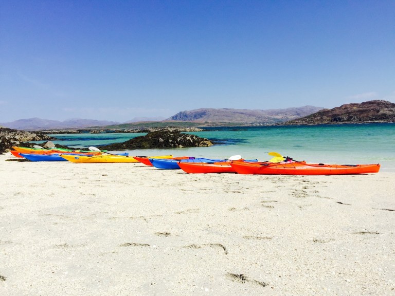 Colorful kayaks lined up on a sandy beach by the sea with rocky hills in the background.
