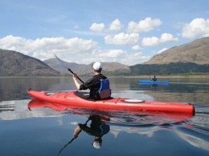Person kayaking on a calm lake with mountains and clouds in the background.