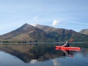 Person kayaking on calm lake with mountains in the background.