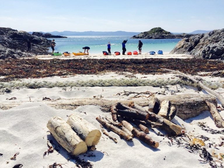 People with kayaks on a sandy beach with seaweed and logs, facing the ocean and distant islands.