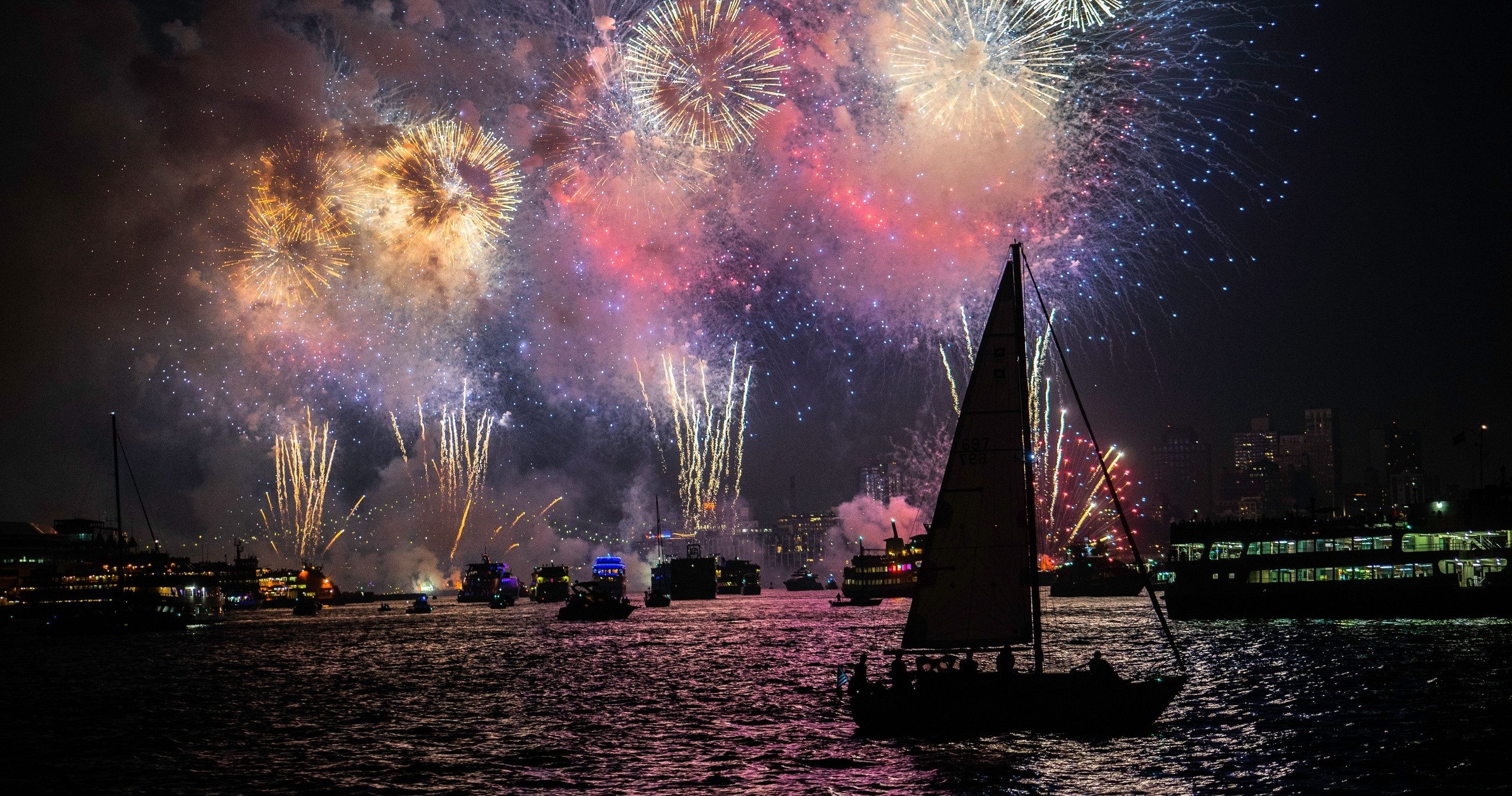 Colorful fireworks over a harbor with silhouetted boats and sailboat at night.