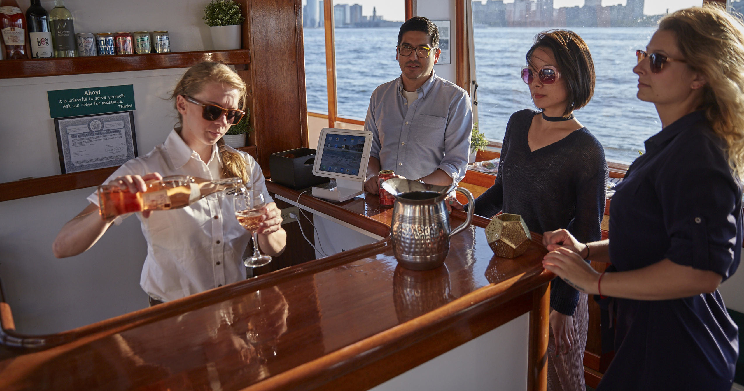 Bartender pours drink for three people on a boat with city skyline in background.