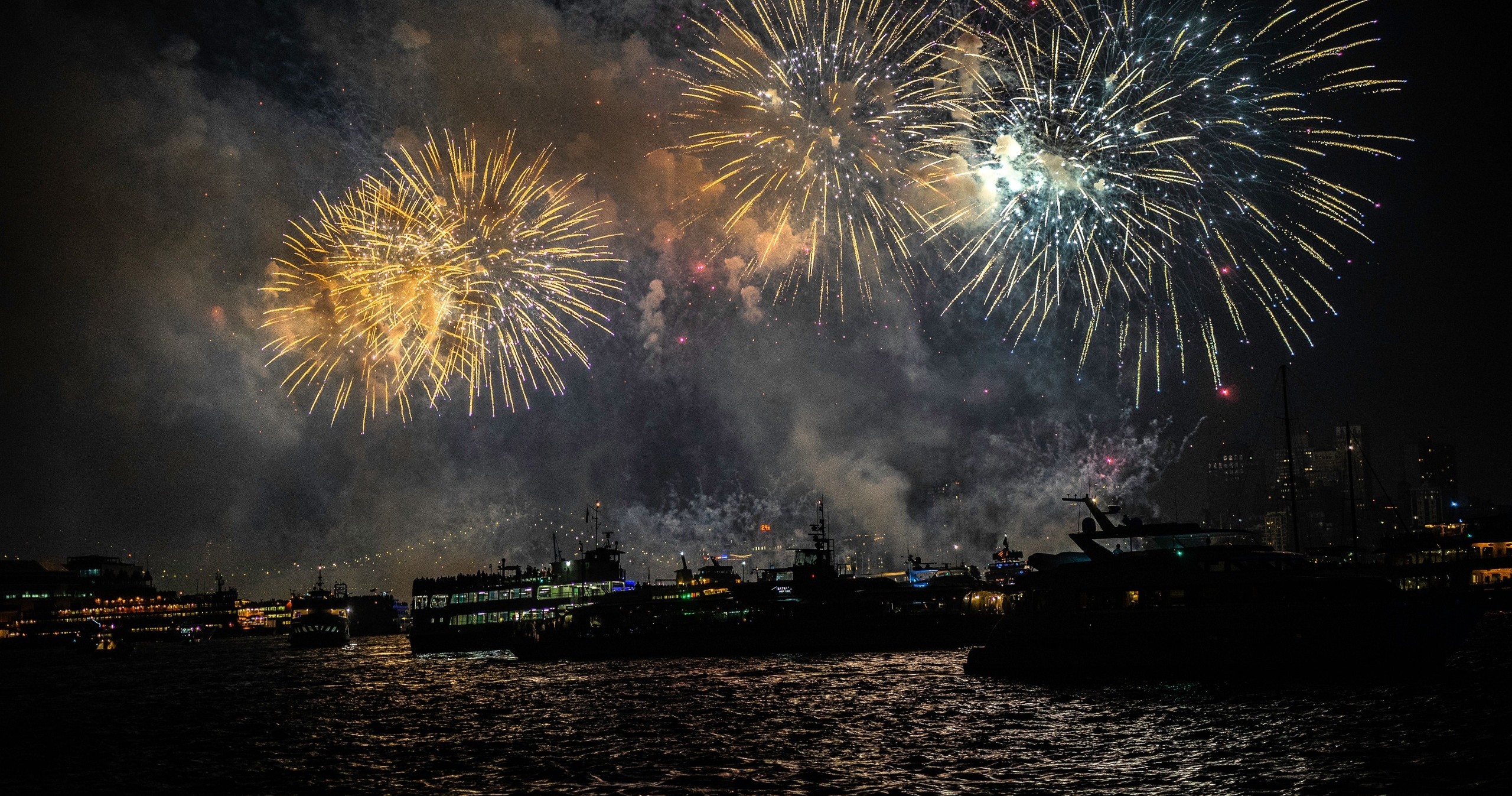 Vibrant fireworks explode over a dark waterfront with silhouetted boats.