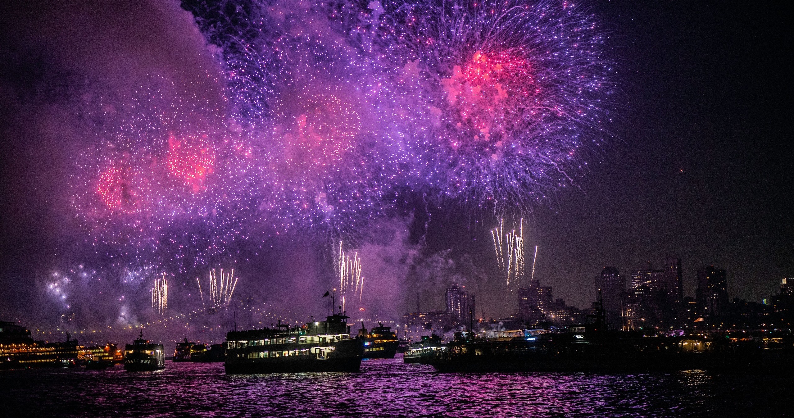 Colorful fireworks over a city skyline at night with boats on the water.