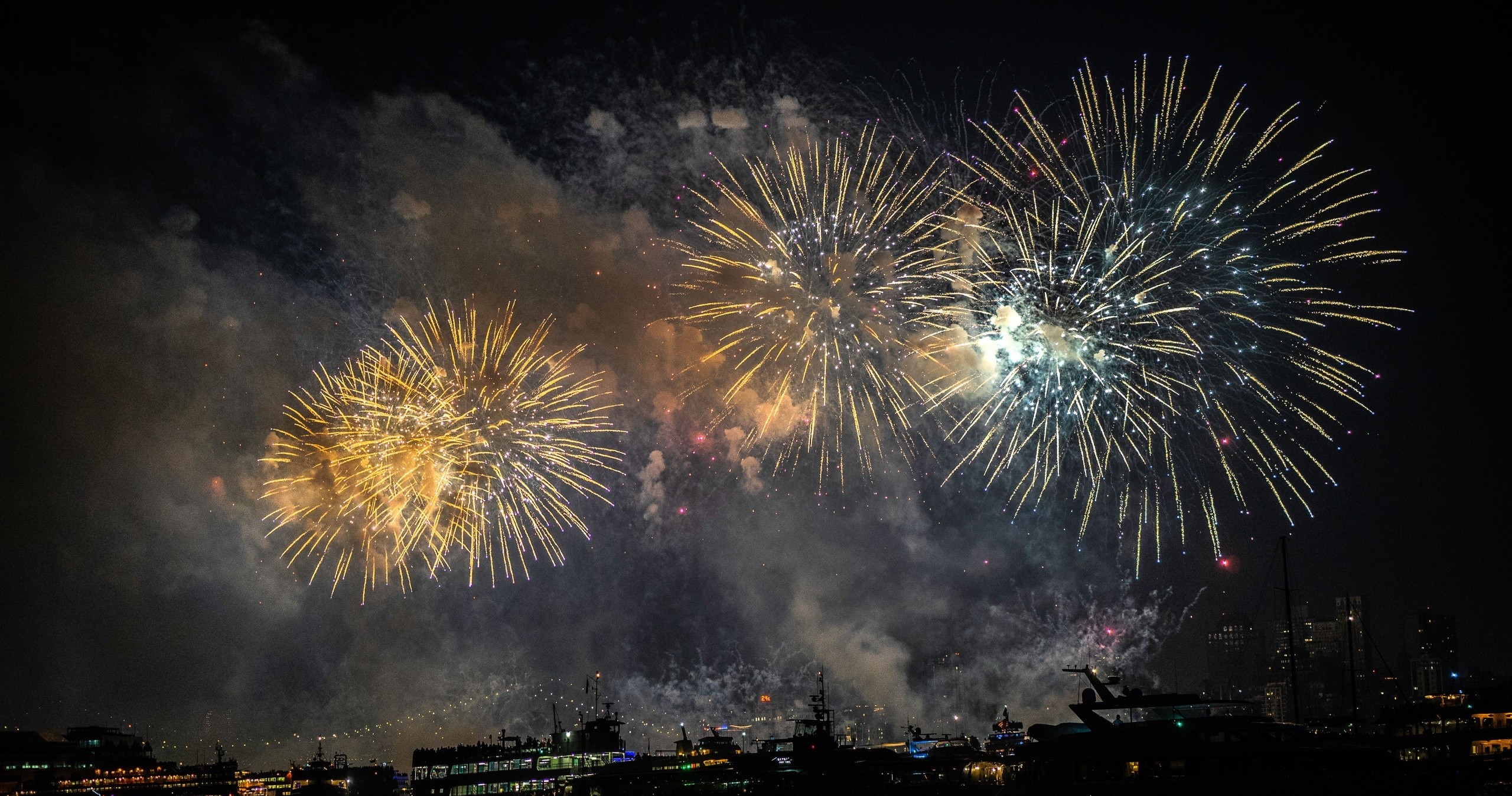 Bright fireworks explode in a night sky over a cityscape with boats.