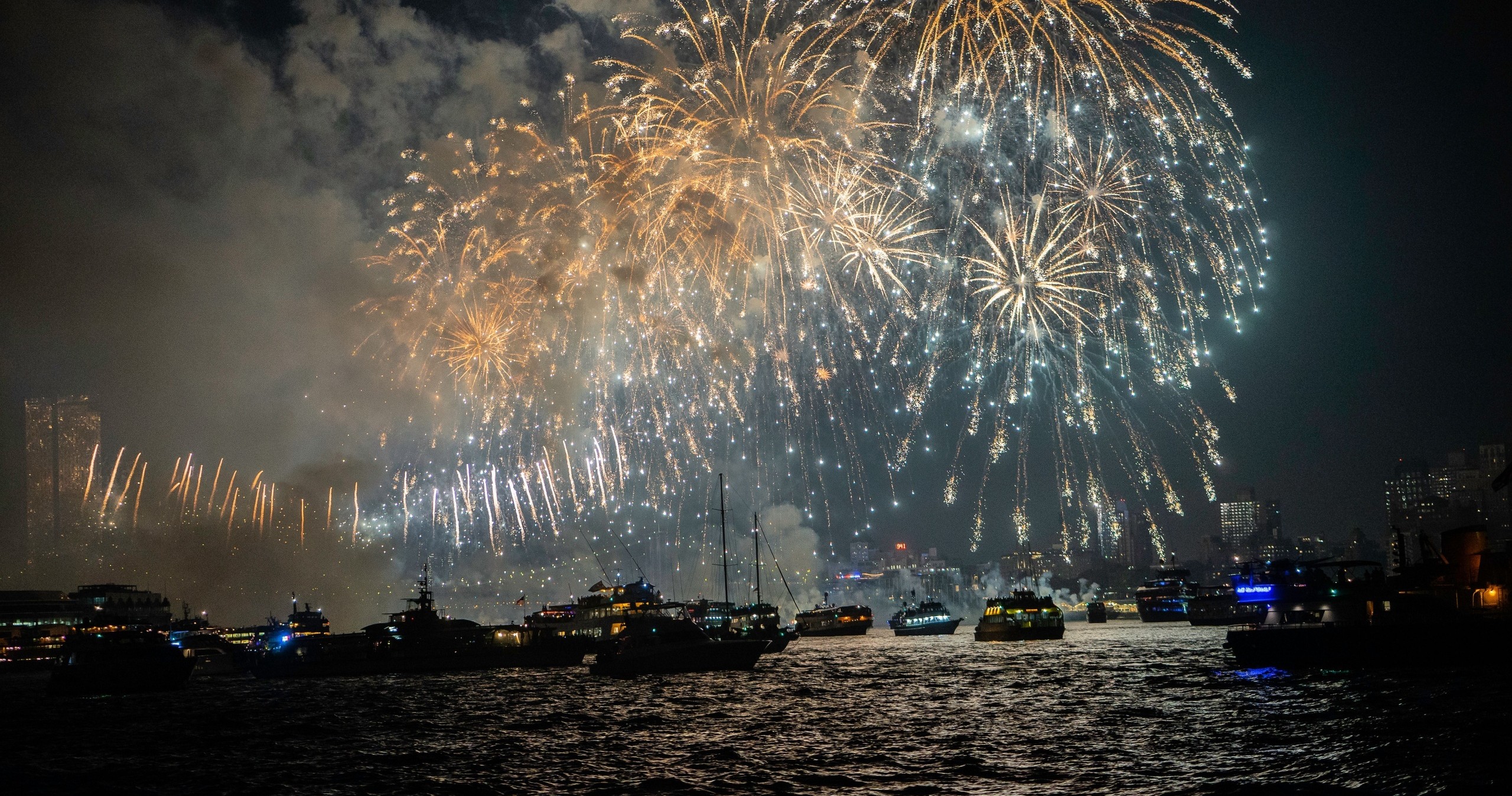 Colorful fireworks light up the night sky over a city and water with boats visible.