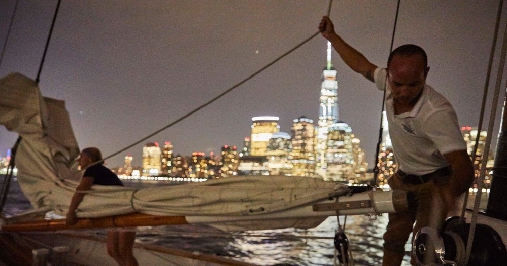 Sailors adjusting sail at night with a city skyline in the background.