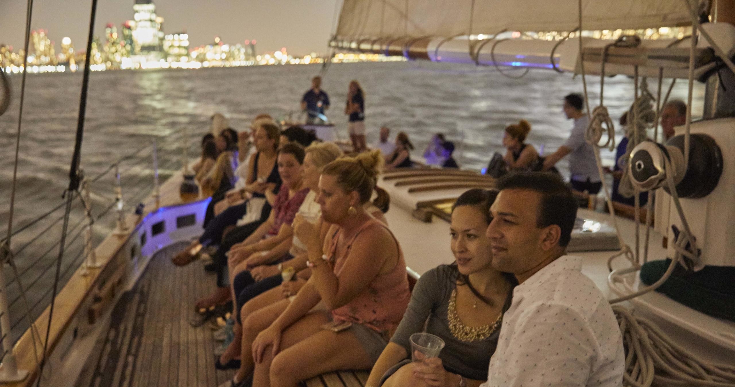 People seated on a sailboat at night with city lights in the background.