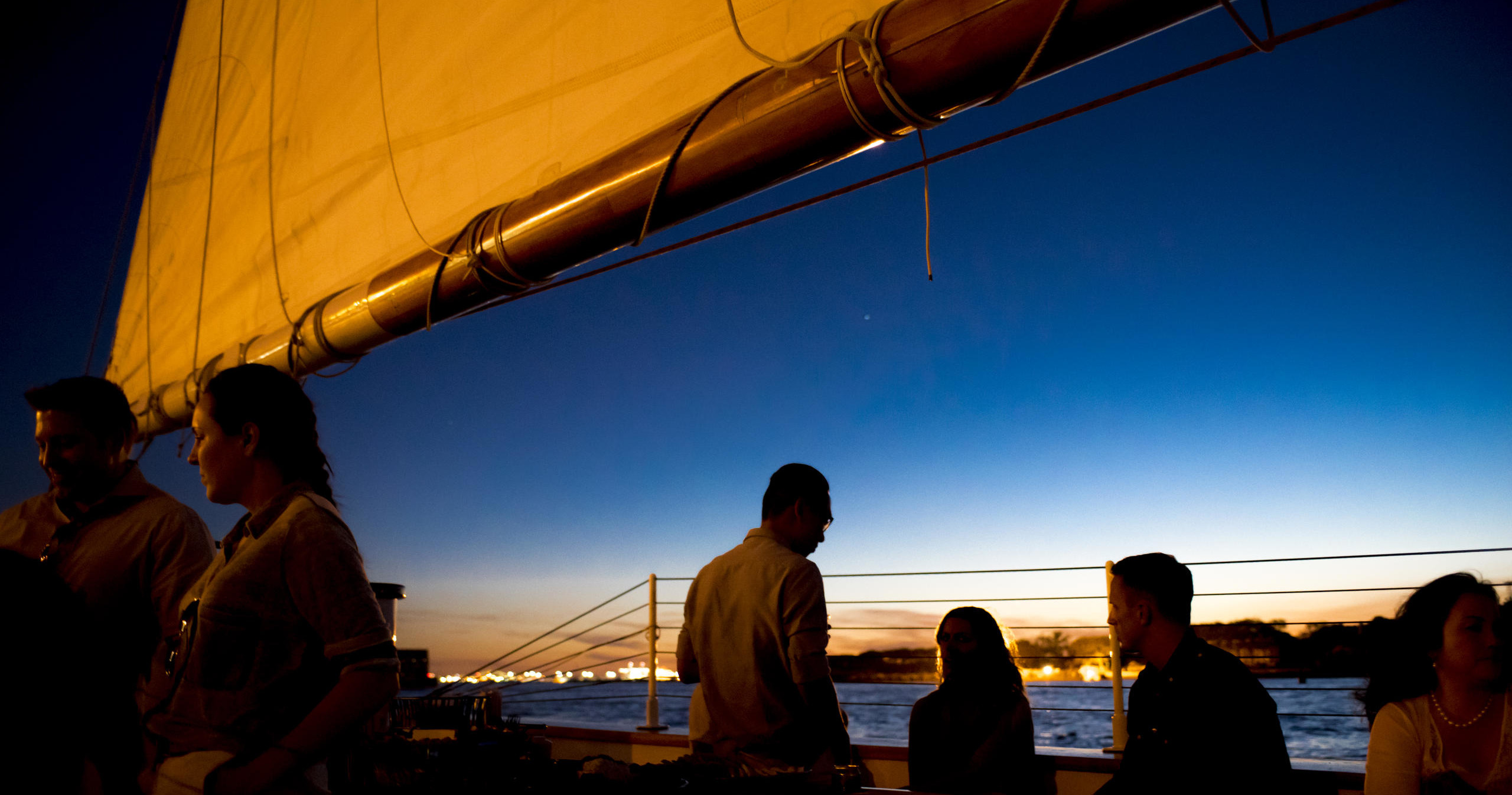 People on a boat at sunset with a sail and water background.