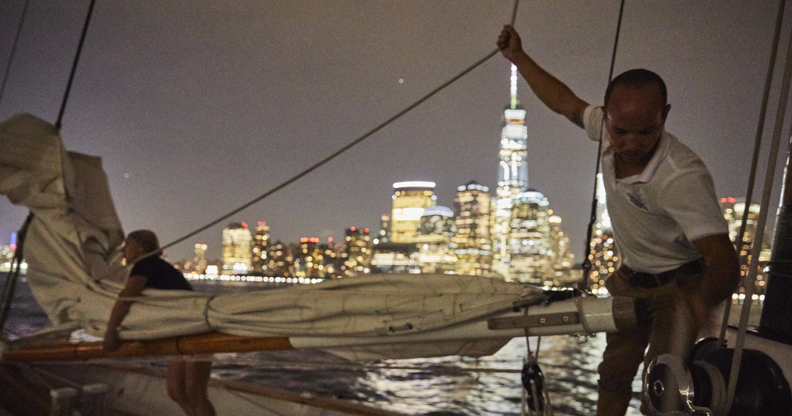 Two people on a sailboat at night with city skyline and lit skyscraper in the background.