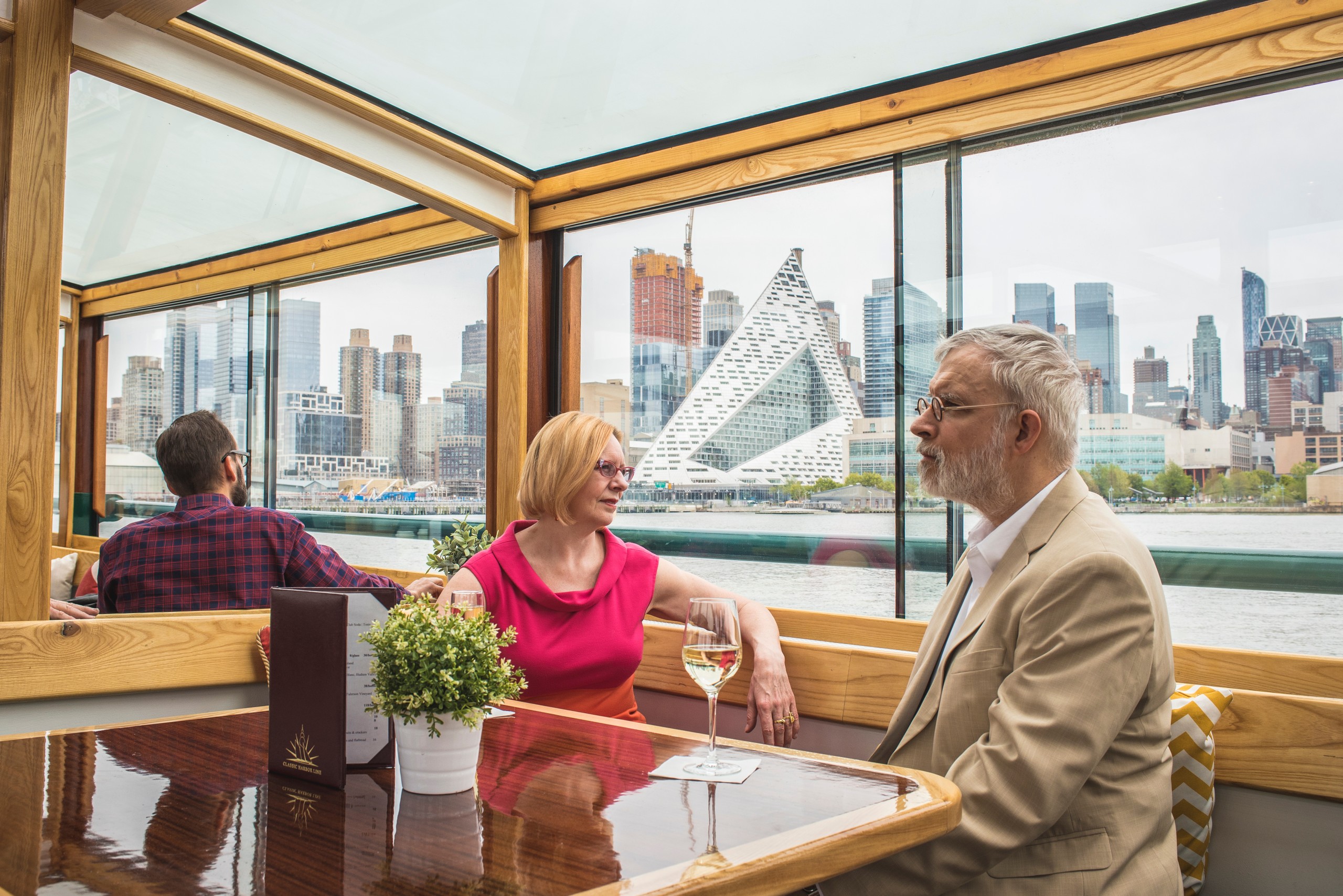 Two people sit at a table on a boat with city skyline in the background.