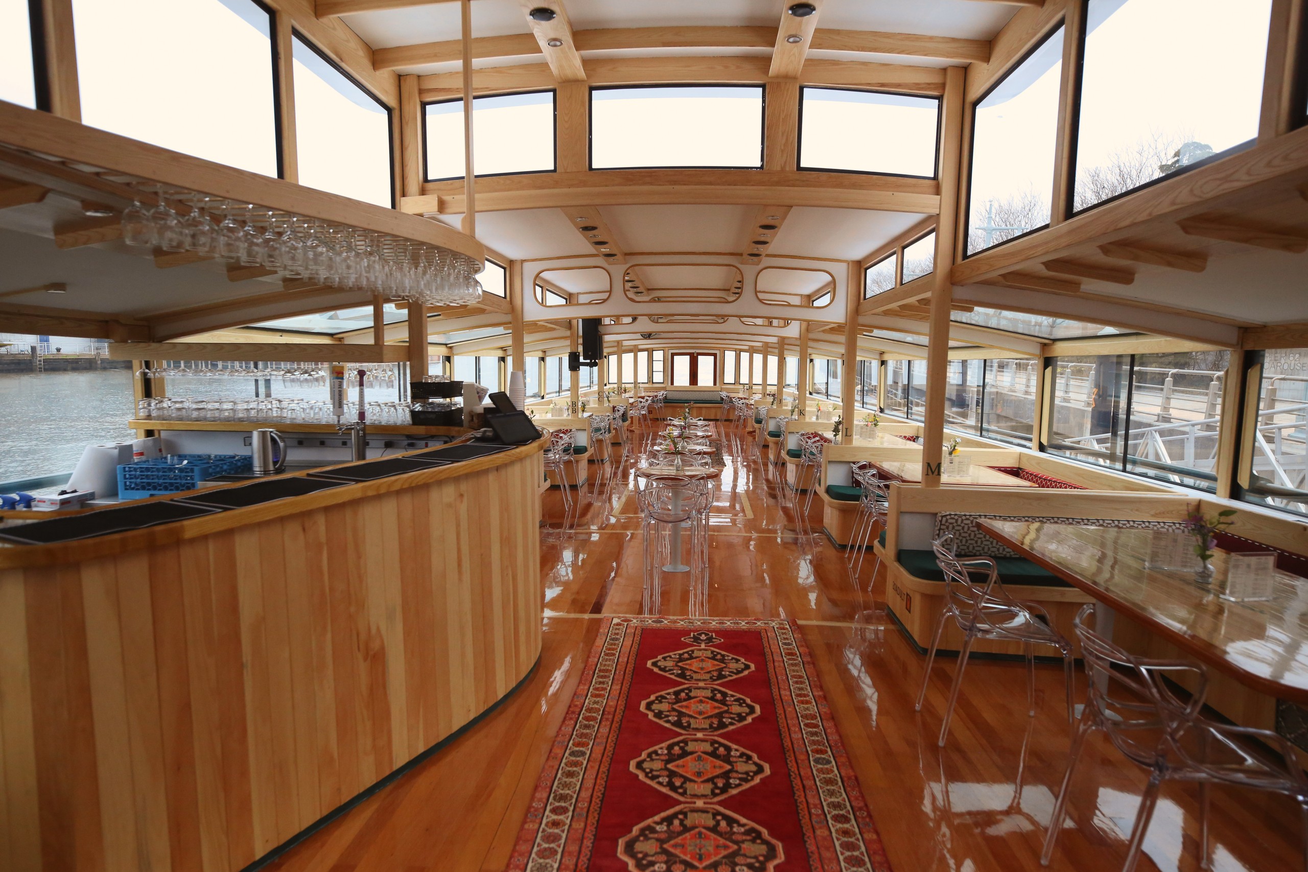 Spacious wooden interior of a boat with large windows and a bar on the left.