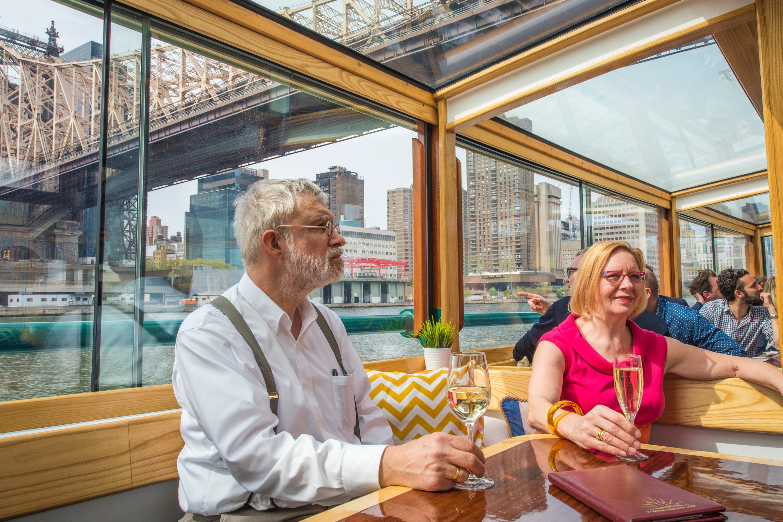 Couple enjoying drinks on a boat, cityscape visible through large windows.