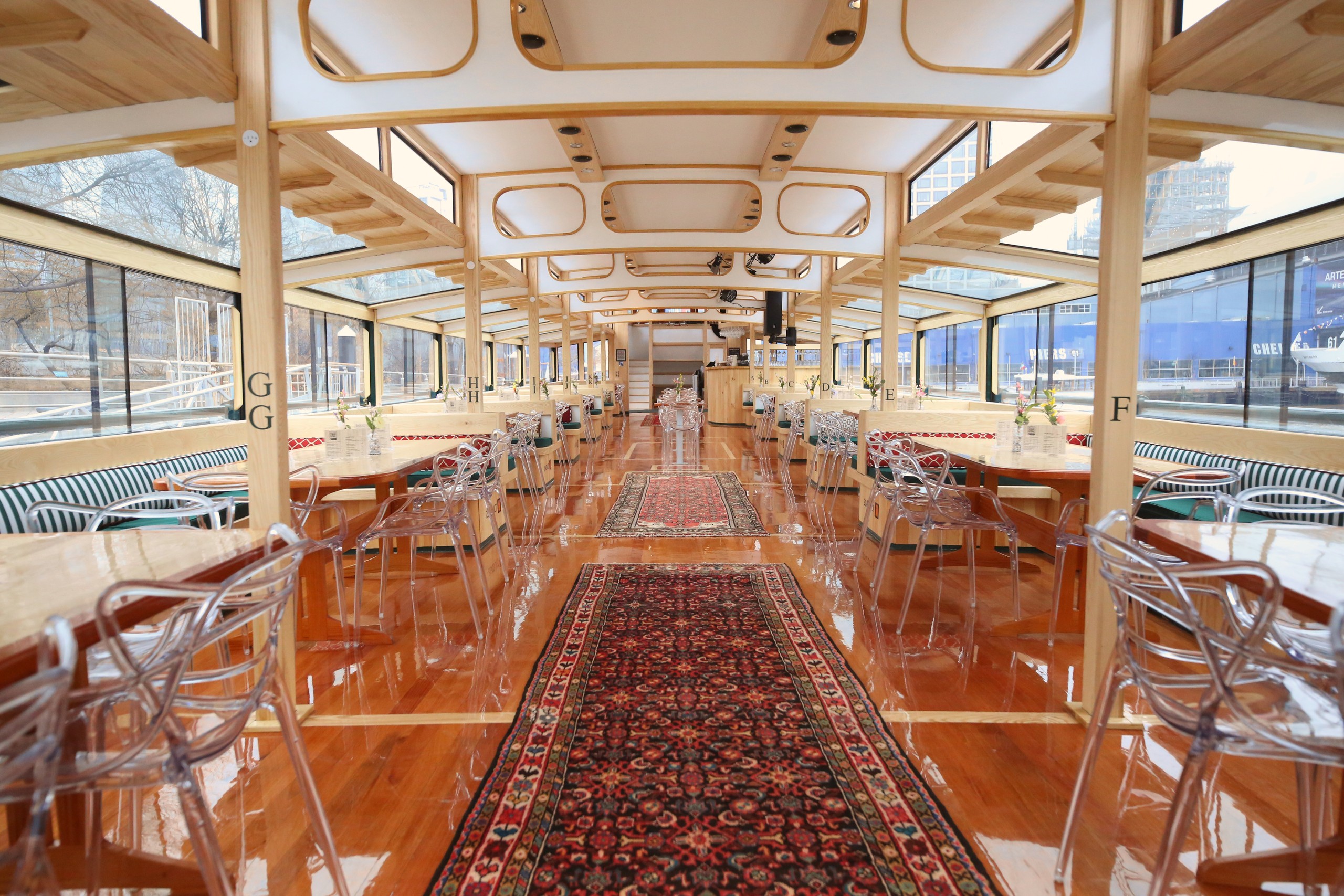 Interior of a boat with clear chairs, wooden floors, and large windows.