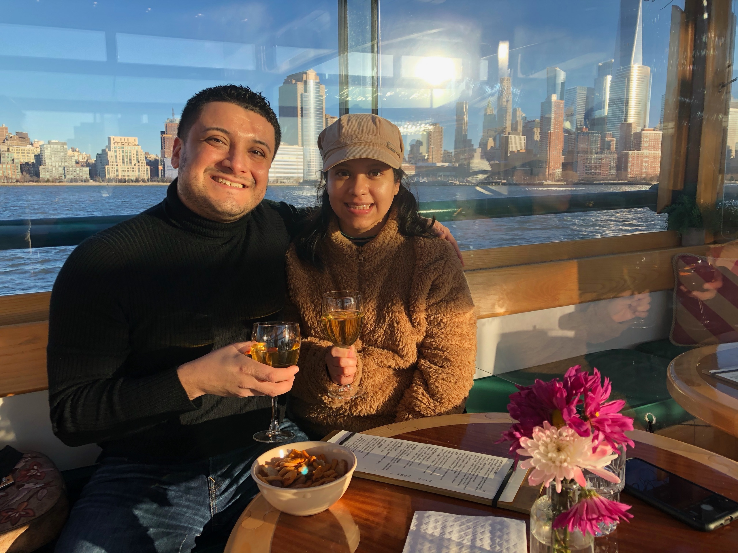 Couple smiling with drinks on a boat, city skyline in the background.