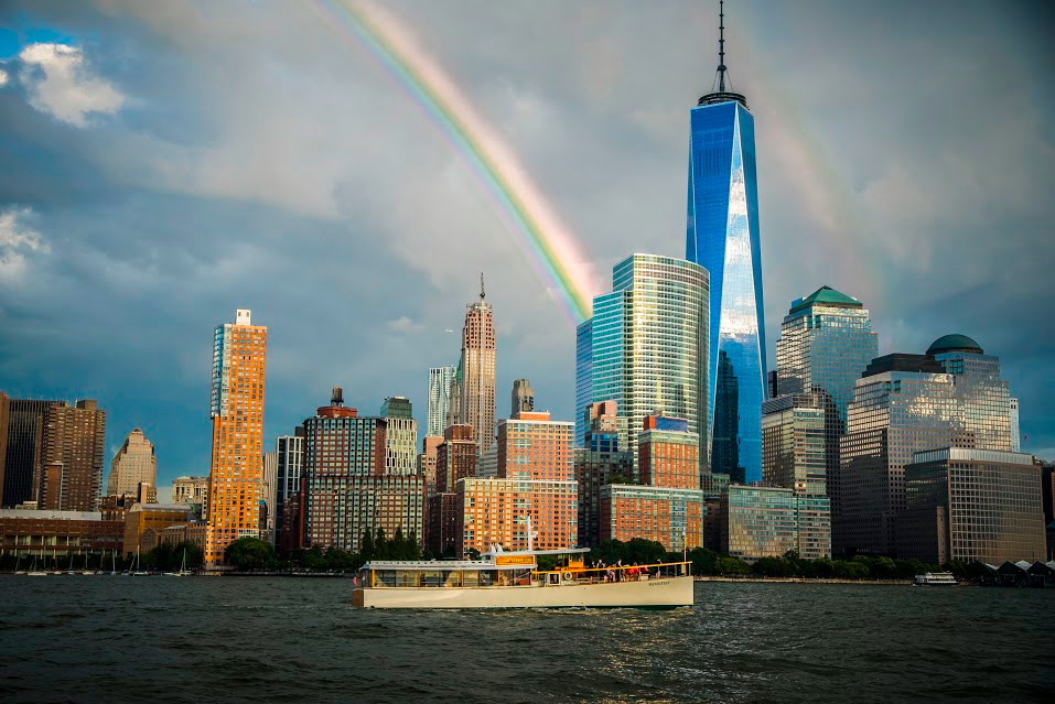 Boat on river, city skyline with tall buildings, and rainbow in cloudy sky.