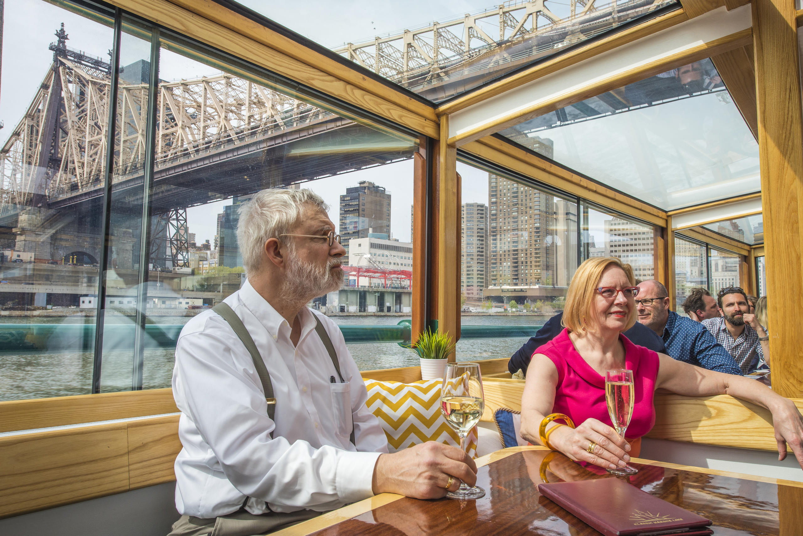 People enjoying drinks on a boat with a city bridge view.