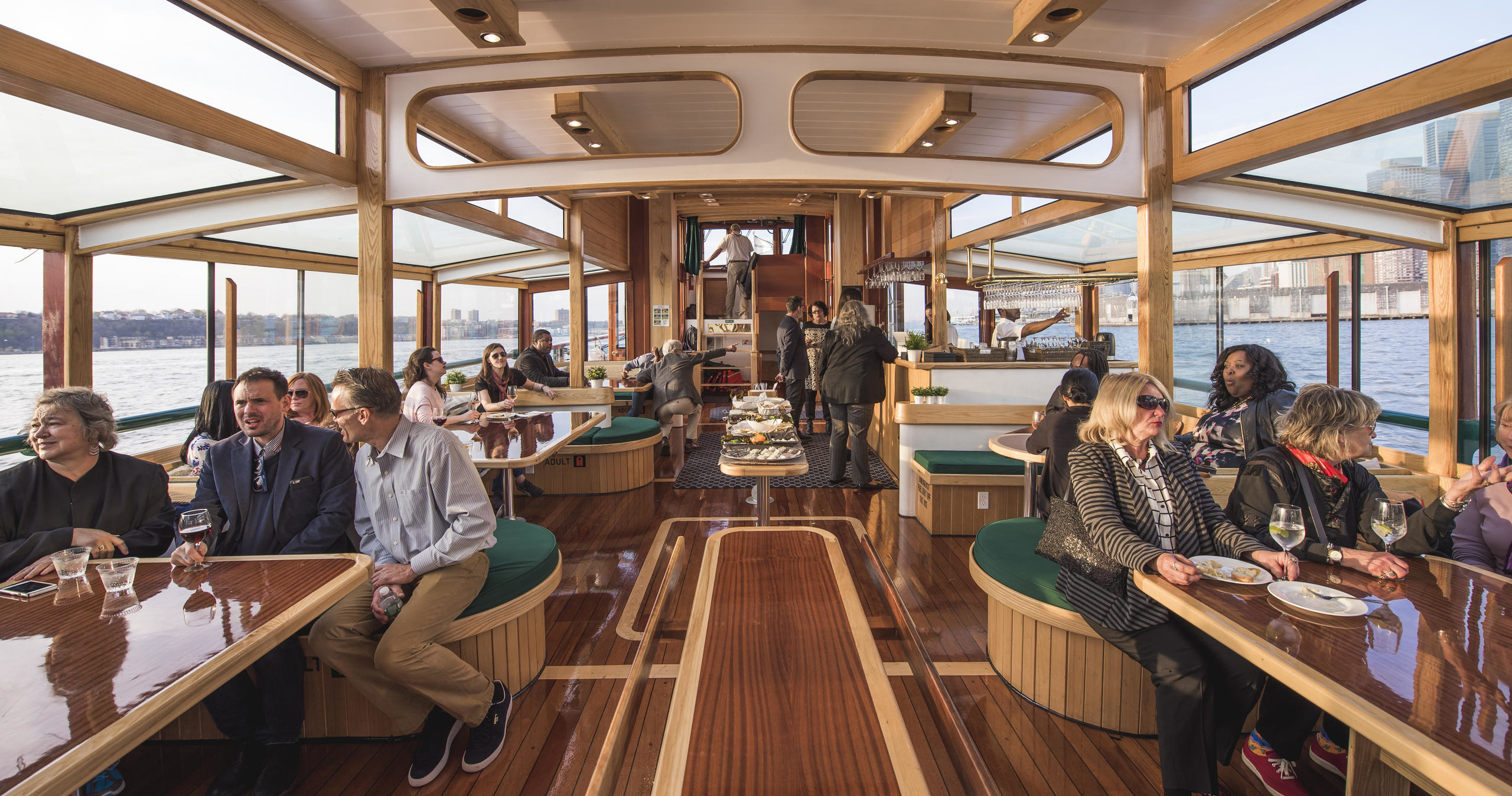People dining on a wooden boat with large windows and a view of water.
