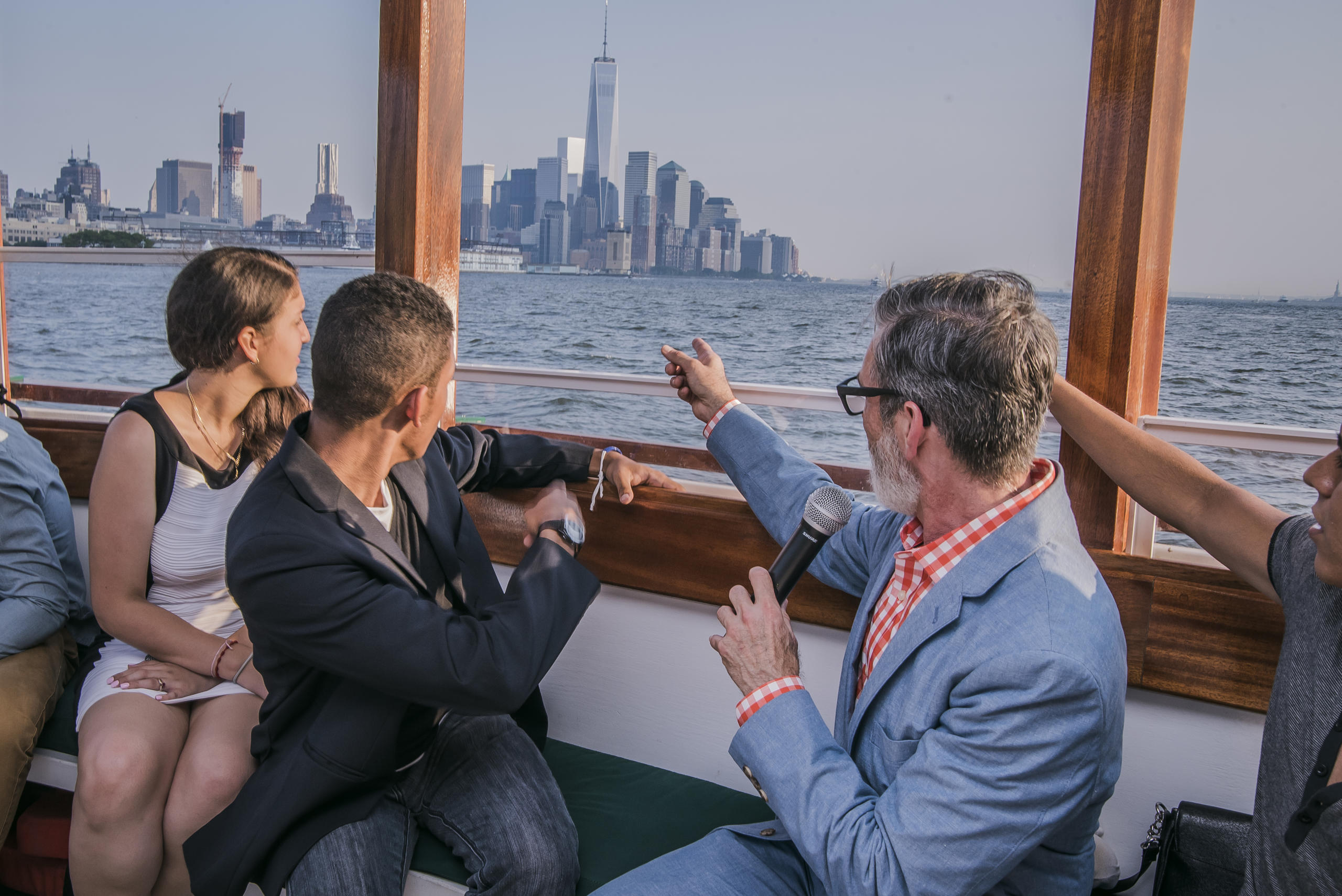 Tour group on boat looks at city skyline, person with microphone points.