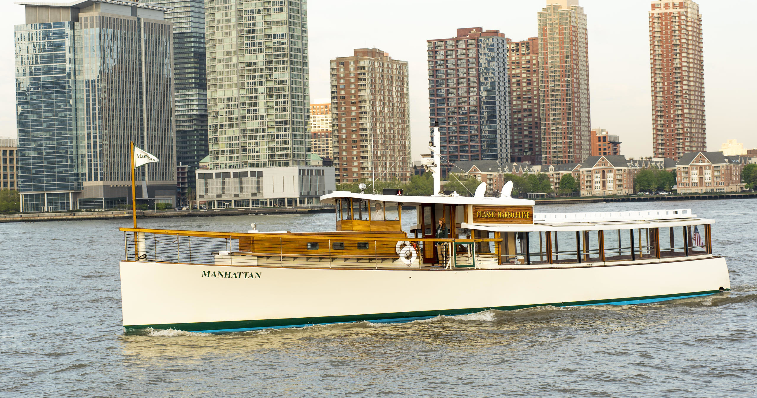 Classic yacht cruising on a river with tall buildings in the background.