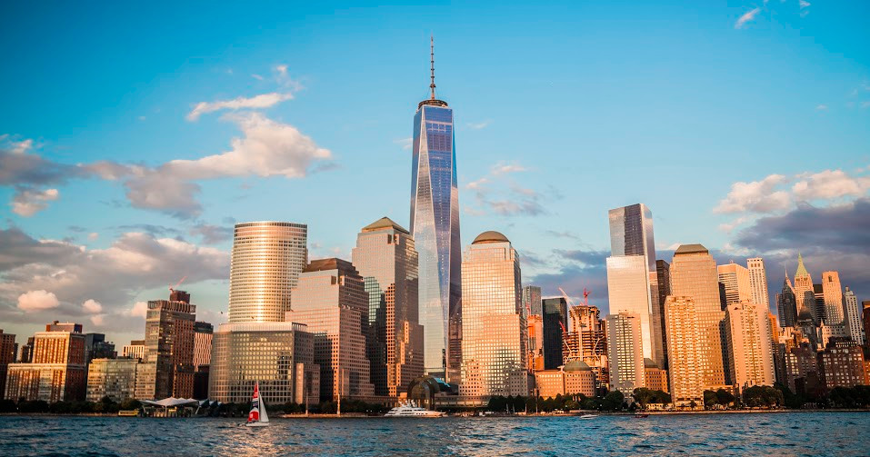 Skyline of tall buildings and One World Trade Center at sunset with water in the foreground.