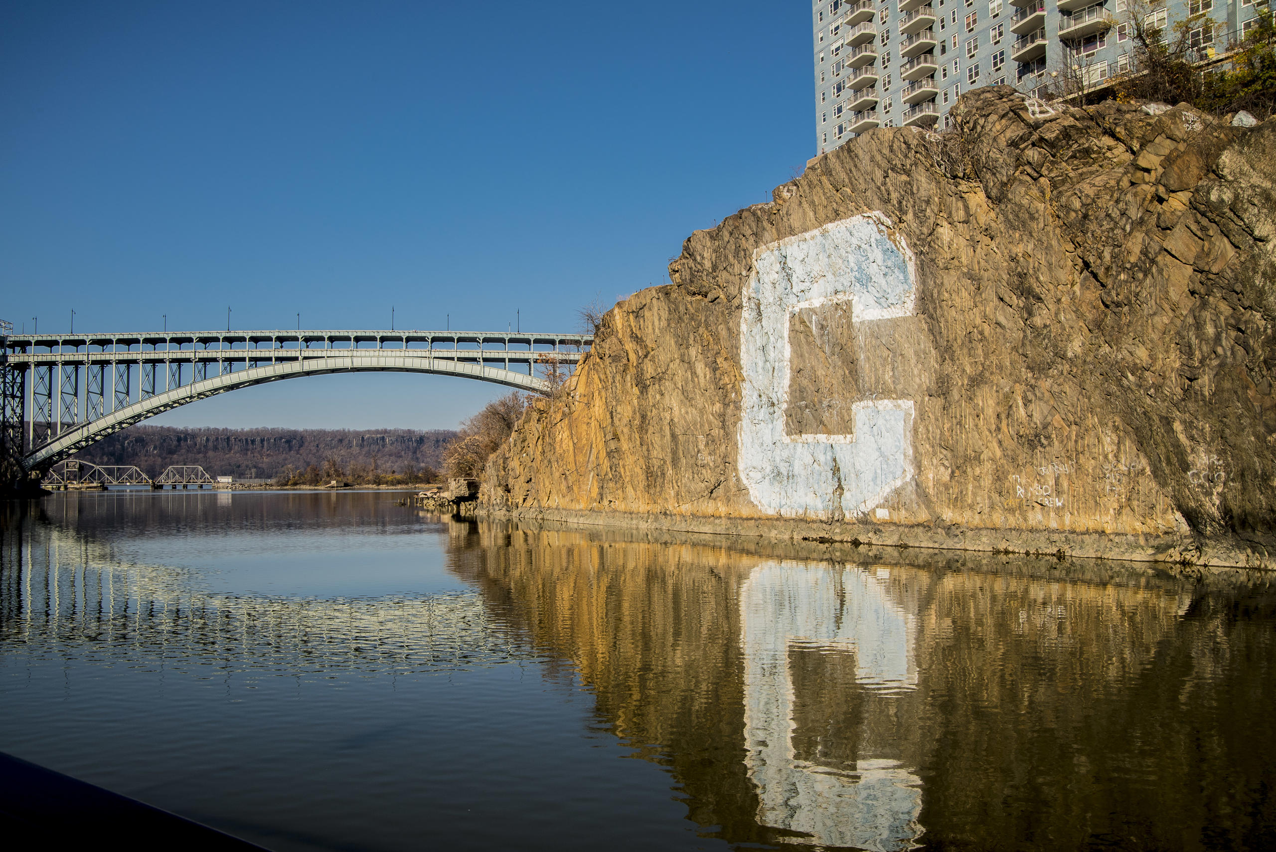 Large letter C painted on cliff by river, with bridge and buildings nearby.