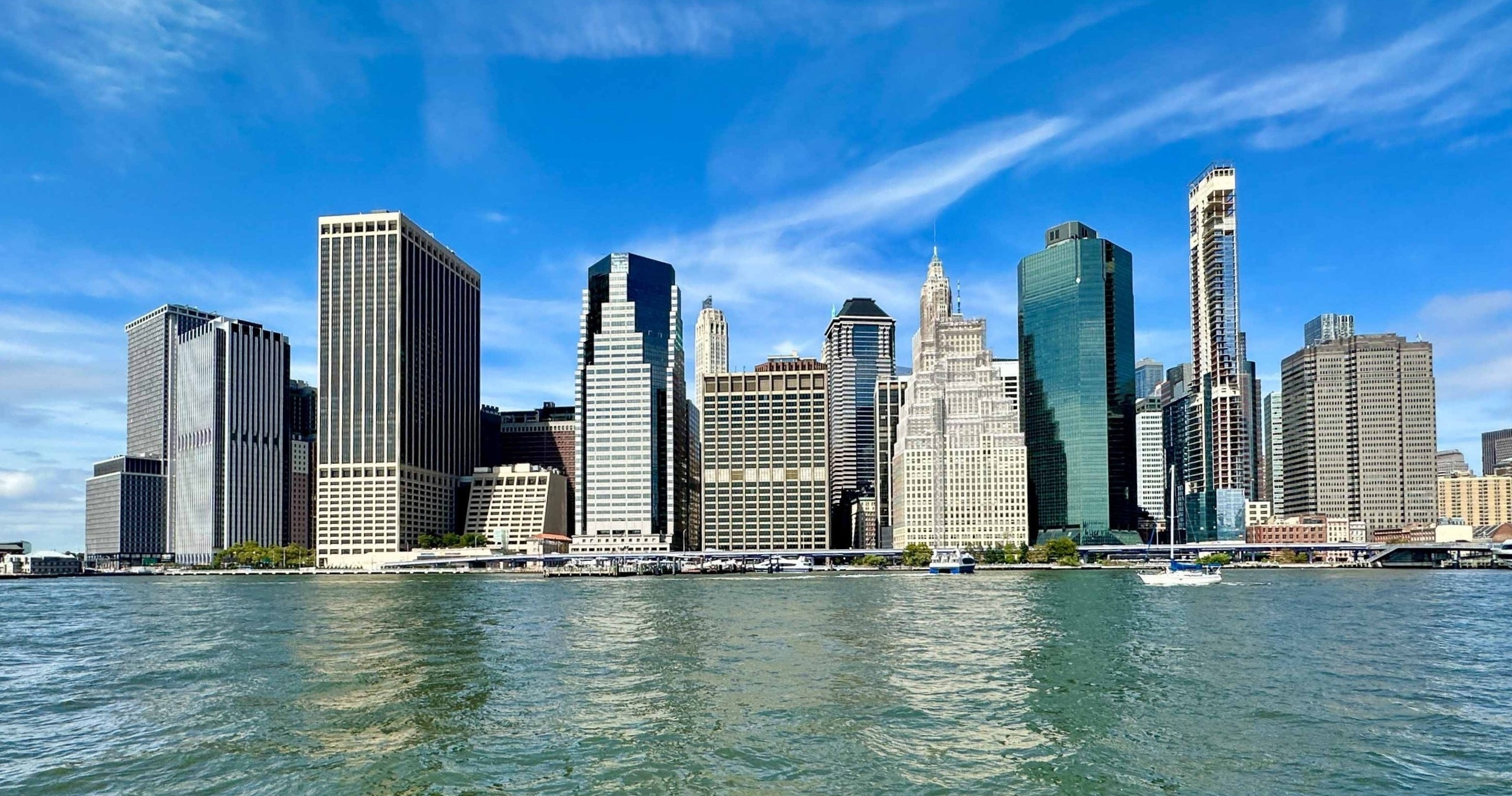 Skyline of tall buildings by a waterfront under a blue sky with clouds.