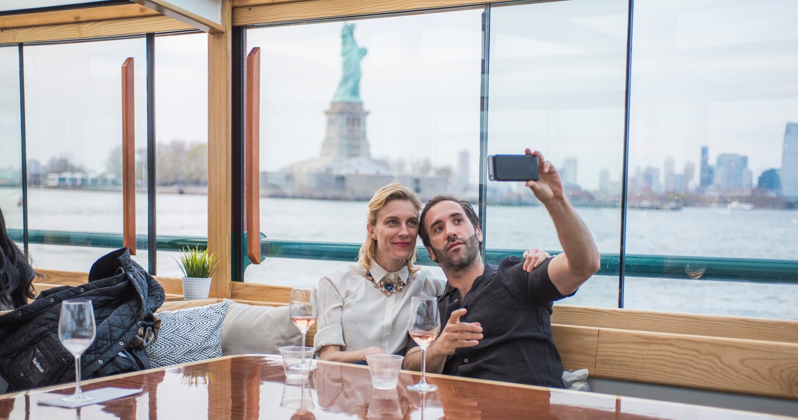 Two people taking a selfie on a boat with the Statue of Liberty in the background.