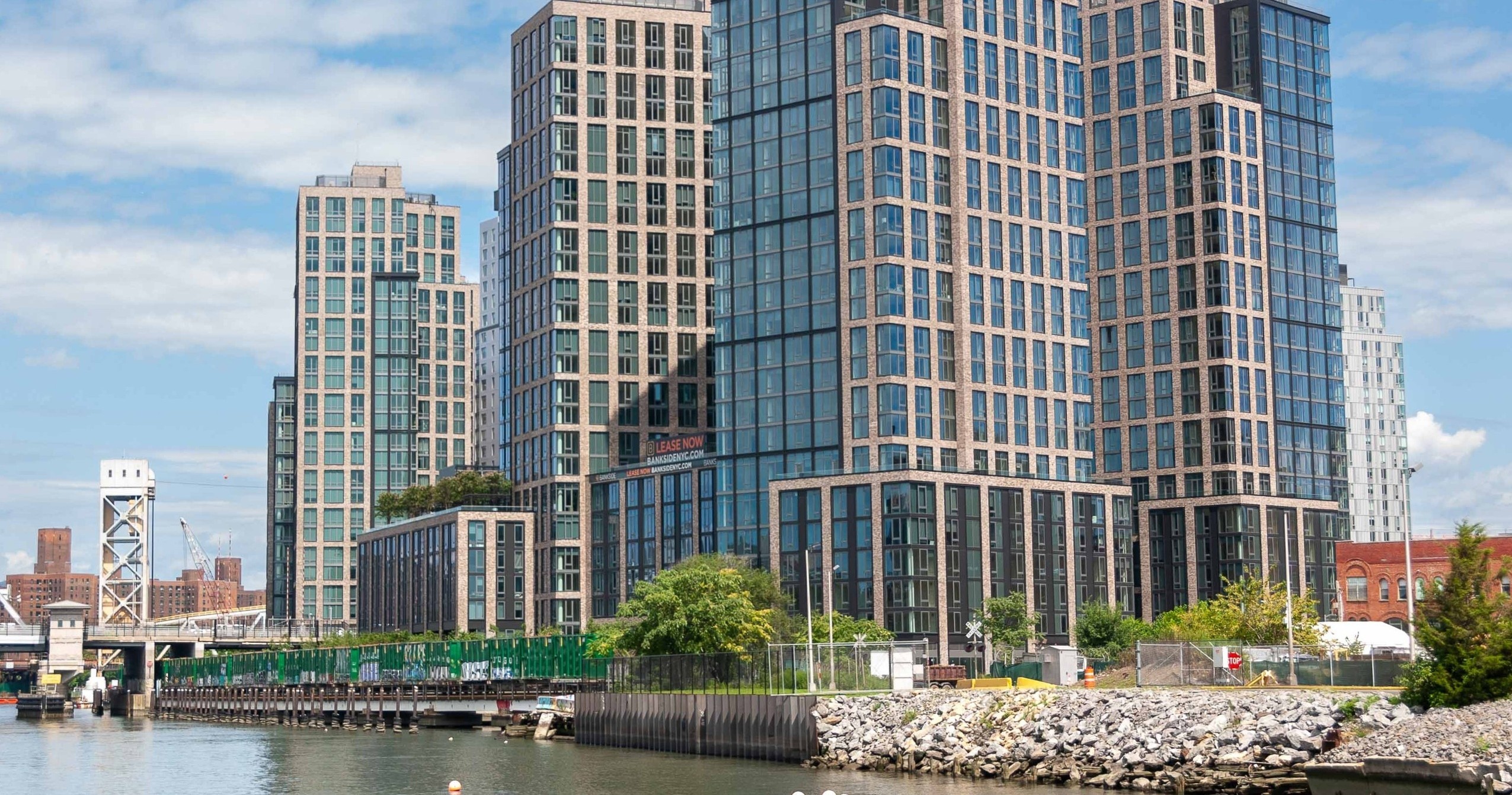 Modern glass skyscrapers by river with bridge and cloudy sky.