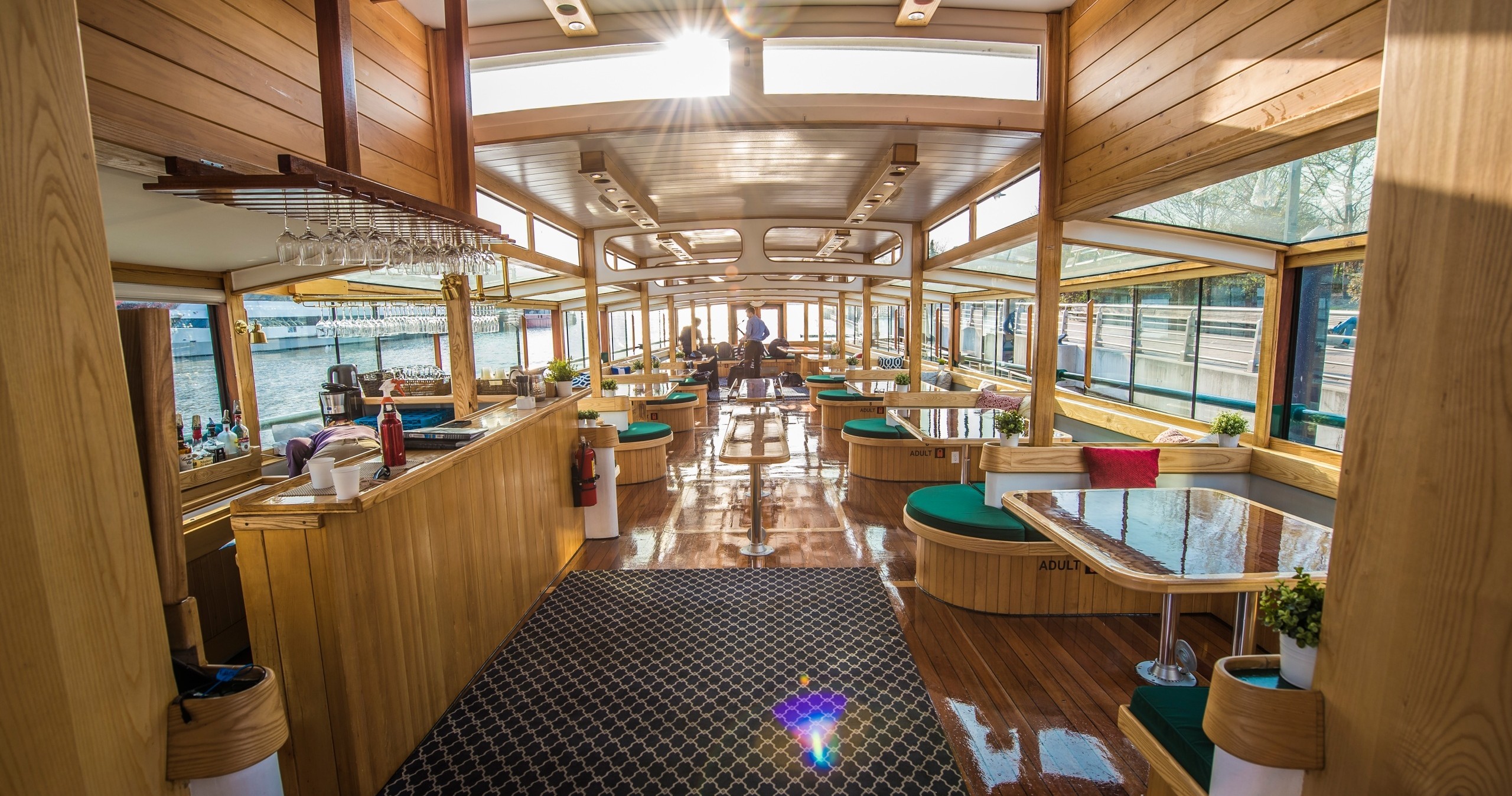 Interior of a sunlit boat with wooden decor, tables, and seating along windows.